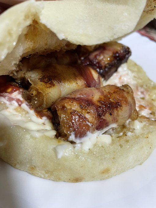 Close-up of a stuffed flatbread or bao bun filled with meat wrapped in bacon and melted cheese, resting on a white plate.