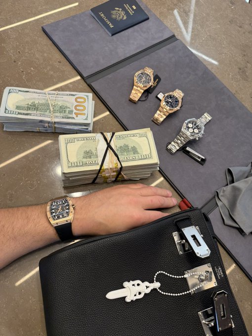 The image shows a wrist adorned with a rectangular gold watch, alongside three luxury watches (two gold and one silver), stacks of $100 bills, a leather document holder, and a black leather case with decorative hardware—all displayed on a polished surface, evoking the high-stakes, flashy aesthetic often associated with crypto wealth culture.