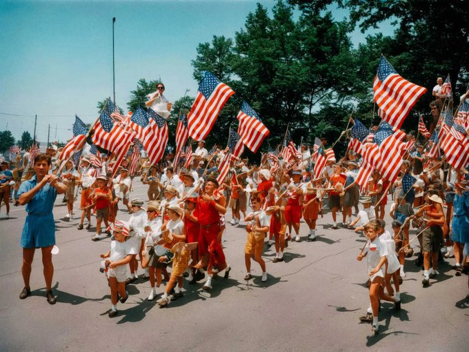 A vibrant photograph of children and adults marching down a street, waving numerous American flags, evoking a patriotic Fourth of July parade.