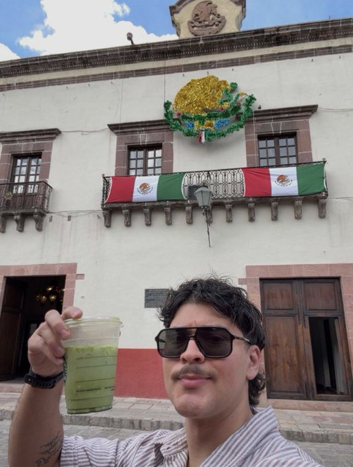 A man with sunglasses takes a selfie in front of a historic building decorated with Mexican flags and a golden eagle emblem, holding a green drink in a plastic cup.