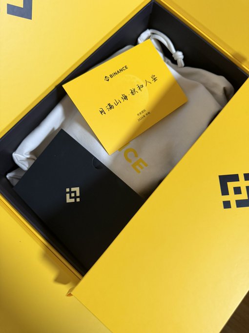 A yellow Binance promotional box containing a black device and a card with Chinese text '前滿山海 敬坤人安'.