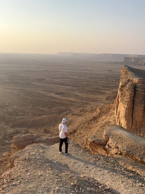 A person in a white shirt and cap stands on a rocky cliff edge overlooking a vast desert landscape at sunset.