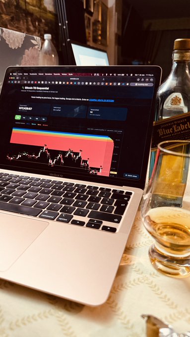 A MacBook displays a bullish Bitcoin/USD price chart alongside a bottle of Johnnie Walker Blue Label and a glass of whiskey on a patterned tablecloth, capturing the lifestyle of a savvy crypto trader taking a celebratory sip. The chart's strong upward momentum visually reinforces the post's "#ZoiTreider" branding, suggesting success and indulgence in the trading world.