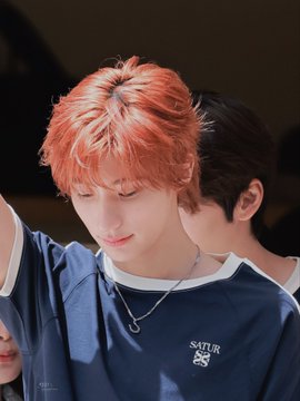 A young man with bright orange hair is looking down, wearing a navy blue jersey that reads "SATUR" with a cross logo, while another person stands close behind him. The image connects to the caption by featuring a member of SATUR, identified as Anton, captured during an event on August 11, 2026.