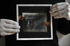 Two hands in white gloves hold up a framed black and white photograph of a young girl's face against a black background.