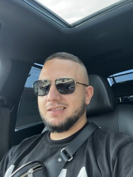 A selfie of a bearded man wearing sunglasses and a black shirt, sitting in the driver's seat of a car with a sunroof visible above.