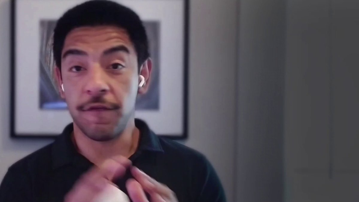 The image features an adult male wearing a black shirt with his hands behind his back as he makes a funny face for the camera. The man appears to be making a funny or silly gesture while looking at the camera, possibly posing for a humorous photo.