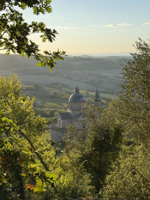 A scenic landscape photograph of an Italian church or monastery with a dome and bell tower, viewed through the branches of trees, with rolling hills and vineyards in the background.