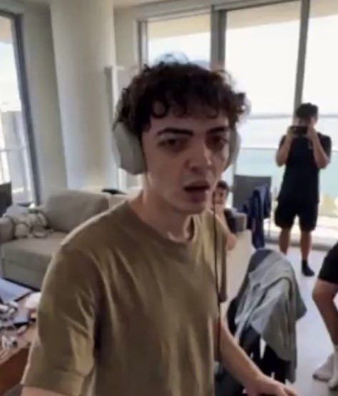 A young man with curly hair wearing headphones and an olive t-shirt sits in a modern apartment while someone behind him films with a phone, illustrating the post's caption about a top SOL trader casually going about their day.