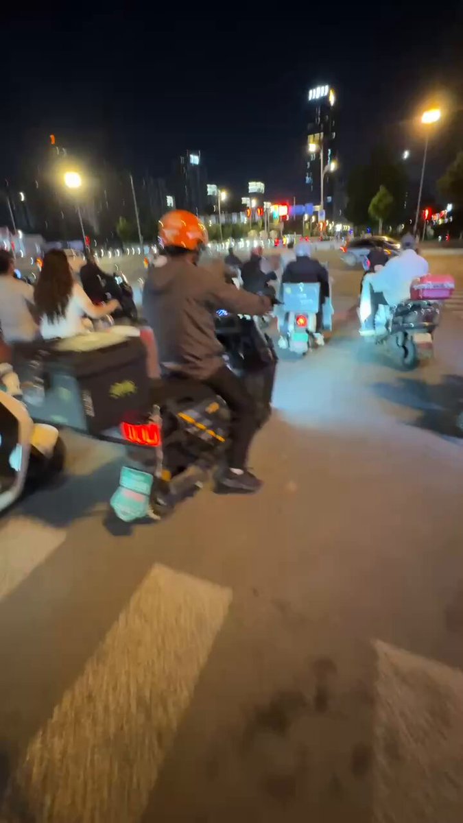 The image shows a group of people riding motorcycles down a street at night, with some parked along the side of the road. The scene appears to be captured from behind one of these riders as they ride by. There are several other motorcycles visible in the background, indicating that this is likely a popular spot for motorcyclists.