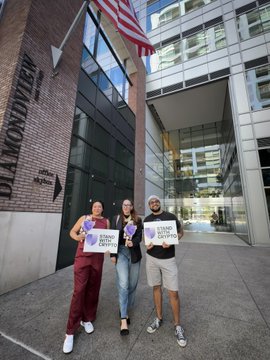 Three people stand on a sidewalk holding protest signs near a building displaying 'NO MONSTERS' and an American flag.