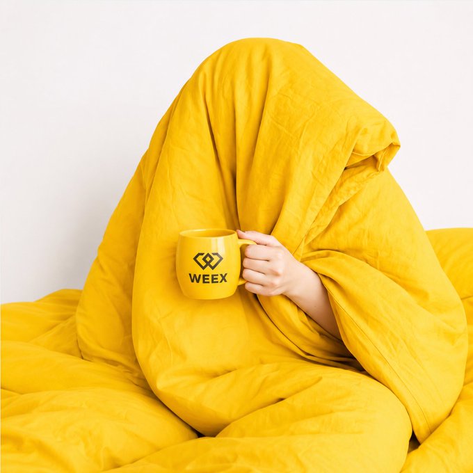 A person completely covered in a bright yellow blanket holds a matching yellow WEEX mug, with only their hand visible.