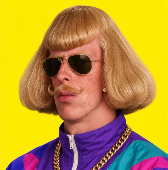 A man with a blonde bowl-cut wig, a curled handlebar mustache, aviator sunglasses, a gold chain, and a purple and teal track jacket poses against a bright yellow background.