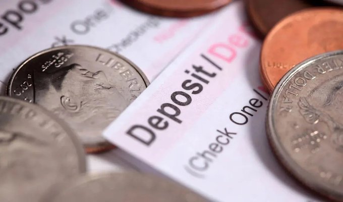 A close-up photograph of various coins scattered around a printed bank statement or receipt that reads 'Deposit / Dep...' and 'Check One/...'