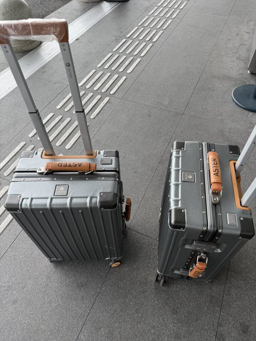 Two silver aluminum suitcases branded with "ASTER" logos stand on a tiled floor, complementing the post's sponsorship shoutout to @Aster_DEX as the traveler heads to Sayram Lake.