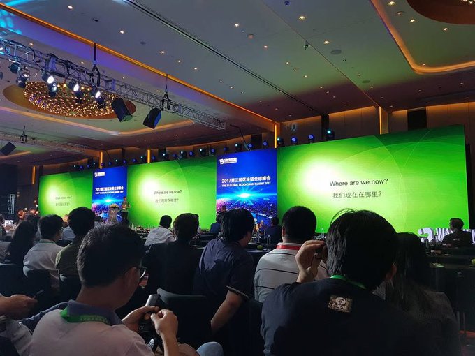 An audience seated at a conference watches large green screens displaying "Where are we now?" with Chinese translations, at what appears to be a tech or industry summit.