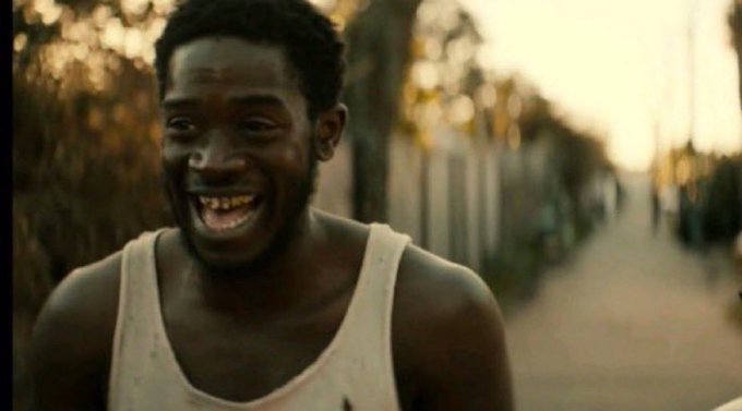 A young Black man smiling broadly, wearing a dirty white tank top on a street with a fence in the background.