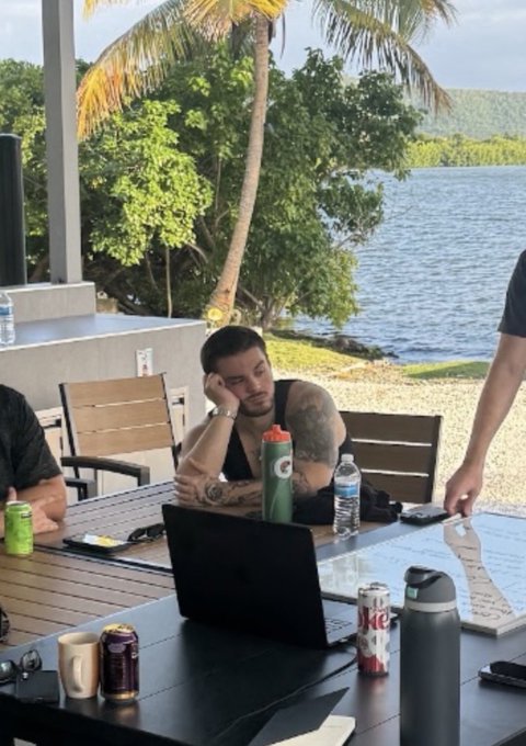 A man sits slumped at an outdoor table with a laptop, surrounded by water bottles, energy drinks, and a soda can, looking dejected while overlooking a serene tropical waterfront. The image humorously captures the post's caption about the disappointment of a Bitcoin investment dropping 50% after buying in at a hyped $125K price point in October.