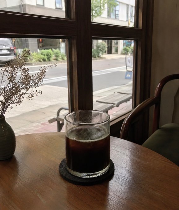 A glass of dark iced coffee sits on a wooden table next to a vase of dried flowers, with a window view of a city street.