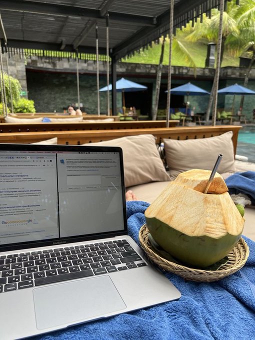 A MacBook Air laptop sits open on a blue towel beside a fresh coconut with a straw in a woven basket, at what appears to be a poolside cabana.
