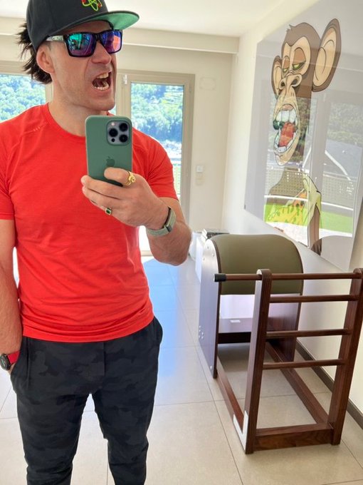 A man wearing a red t-shirt, sunglasses, and a cap yells excitedly while taking a selfie with his phone in front of a large cartoon monkey painting.