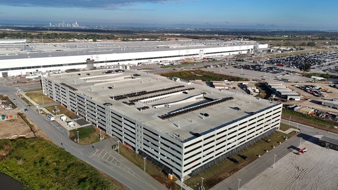 Aerial view of a large Tesla Gigafactory manufacturing complex with multiple parking lots full of vehicles and the city skyline visible in the distance.