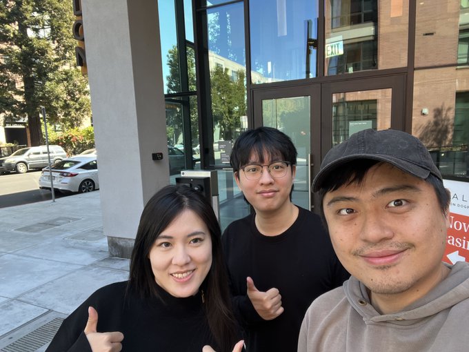 A selfie of three smiling young people (two giving thumbs up) standing outside a building with large glass windows and an EXIT sign visible.