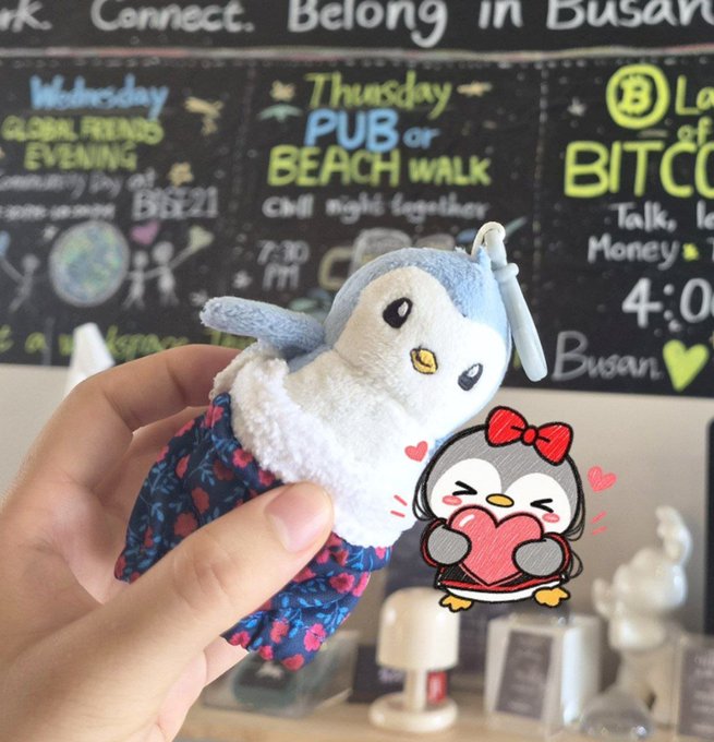 A hand holds a small plush penguin toy wearing a blue dress in front of a chalkboard menu featuring events like 'Wednesday GLOBAL FRIENDS EVENING' and 'Thursday PUB or BEACH WALK' in Busan, with a cartoon penguin sticker added.