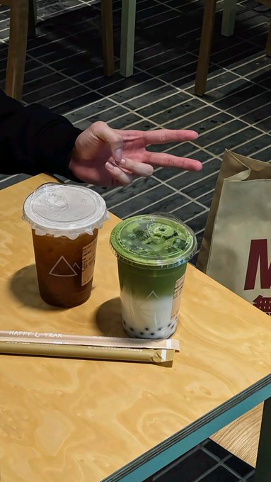 Two drinks from a café sit on a wooden table, one iced tea and one matcha latte with boba, alongside a paper bag featuring a large "M" logo. A person flashes a peace sign in the background, mirroring the post's theme of finding value in quiet, underappreciated work rather than the loudest projects in the crypto space.