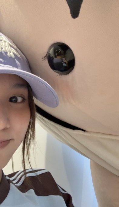 A selfie of a person wearing a purple and white cap and a striped shirt, posing next to a large plush character.