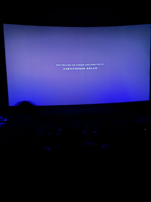 A photograph taken inside a dark cinema showing a glowing blue screen with the text 'WRITTEN FOR THE SCREEN AND DIRECTED BY CHRISTOPHER NOLAN'.