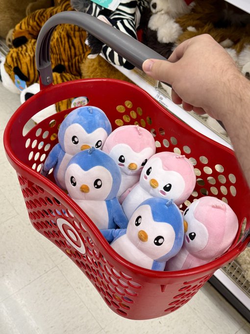 A hand reaches down to grasp the handle of a red Target shopping basket filled with blue and pink Pudgy Penguins plush toys, with additional plushies visible on store shelves in the background.