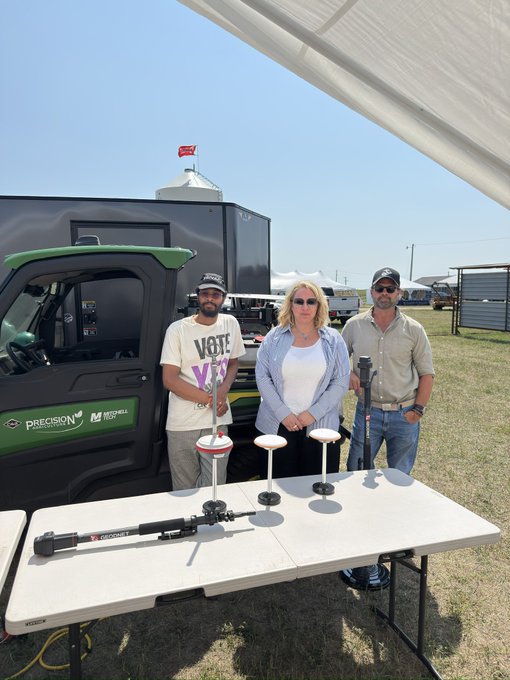 Three people standing behind a table displaying agriculture equipment at an outdoor event, with a trailer labeled 'Precision' and 'Mitsubishi' in the background.