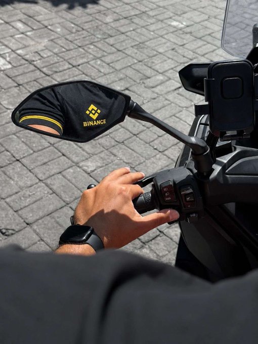 A motorcyclist grips the handlebar with a hand wearing a smartwatch, while a side mirror prominently displays the "BINANCE" logo with its yellow emblem, paired with a mounted phone—illustrating a focused ride free of distractions, just as the caption urges "Eyes strictly ahead."