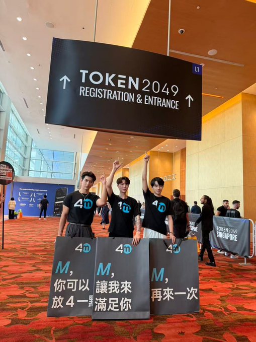 Three young men wearing black "40" t-shirts pose under a "TOKEN 2049 REGISTRATION & ENTRANCE" sign at the Singapore crypto conference, holding signs with Chinese text that translates to "M, you can let me get 4 satisfaction again." The image illustrates the post's warning about the 4M exchange, with attendees displaying promotional "40" branding at the very event where the exchange was reportedly being used to attract users before its suspected exit scam.