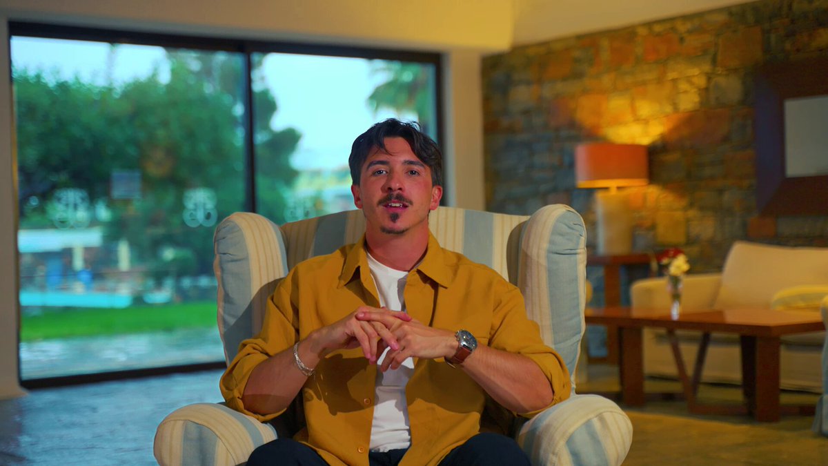 The image features a man sitting comfortably in an armchair, wearing a yellow shirt. He appears to be posing for a photo or recording a video. The background of the image shows a living room with a couch and a dining table. There is also a window visible in the scene that provides natural light into the space.
