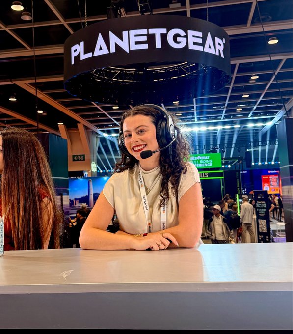 A smiling woman wearing a headset sits at a booth beneath a circular 'PLANETGEAR' sign at what appears to be a gaming or electronics convention.