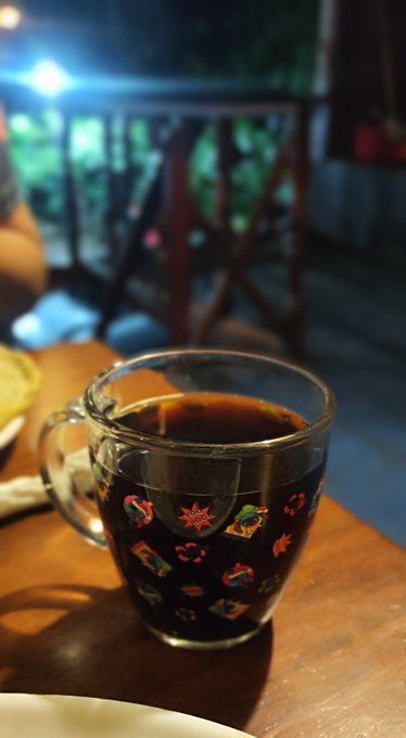 A clear glass mug decorated with colorful festive patterns sits on a wooden table, filled with a dark liquid and featuring a tea bag steeping inside.