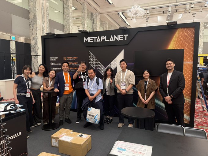 A group photo of people posing at a 'METAPLANET' booth at a conference, with sponsor logos including Bitcoin, Blockchain, and Bitcoinpoint visible on the backdrop, and some attendees holding Web3 bags.