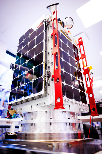 A small satellite with solar panels and red structural framework sits on a cylindrical metallic base, featuring the text "ZEPHYR" on its side.