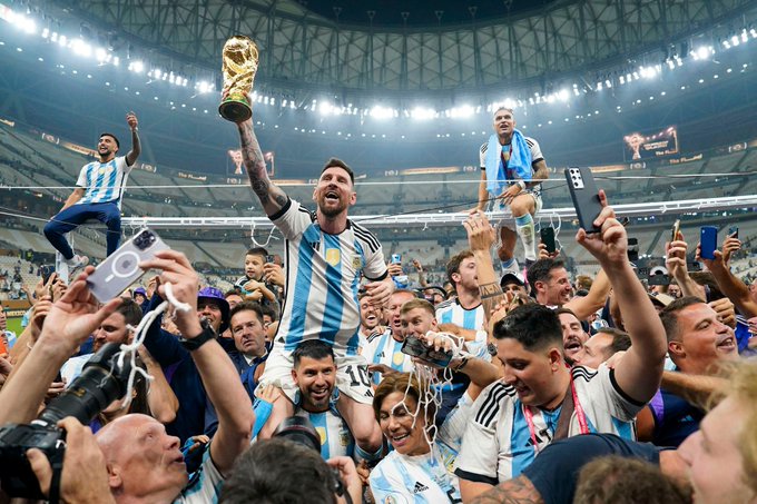 Lionel Messi holds up the FIFA World Cup trophy in an Argentina jersey, surrounded by celebrating teammates and fans inside a stadium.