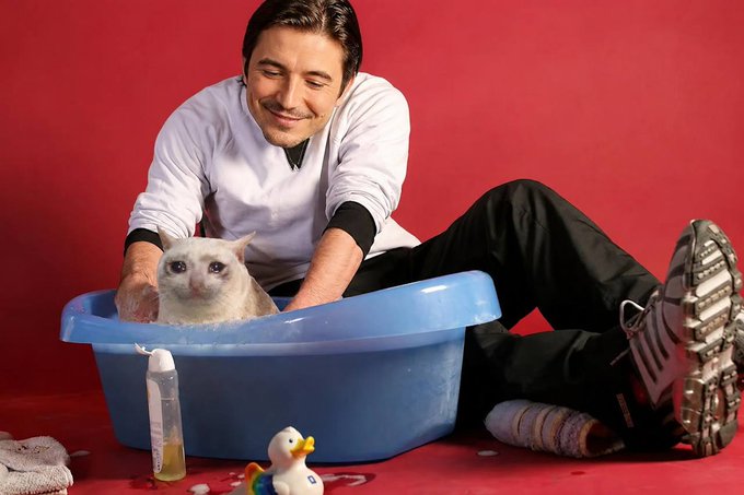 A man sits on the floor giving his cream-colored cat a bath in a blue plastic tub against a red backdrop, with a rubber duck, shampoo bottle, and rolled towel nearby—capturing the humorous contradiction of choosing between typical masculine and caring, nurturing activities, perfectly illustrating "Why not both?"