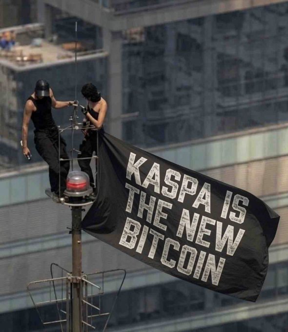 Two people in black attire hang a large banner reading "KASPA IS THE NEW BITCOIN" from a beacon tower atop a building, promoting a crypto promotional stunt. The image relates to the post's hopeful investment in Kaspa for the future, reflecting the bold ambition of those betting on the cryptocurrency's long-term value.