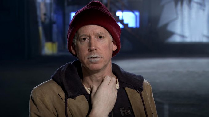 A man wearing a red beanie and a brown jacket over a dark hoodie, standing in what appears to be an industrial or warehouse setting with a surprised expression and his hand near his chin.