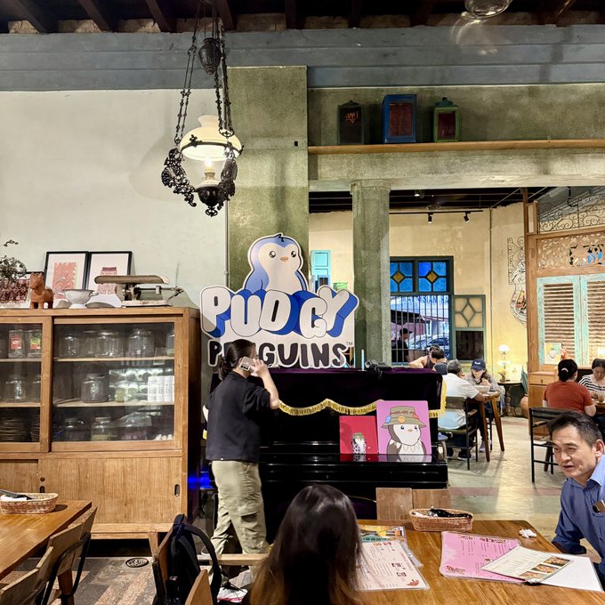 A vibrant interior of a café or restaurant featuring a 'Pudgy Penguins' logo display, with patrons dining at wooden tables and a hanging ornate lamp.