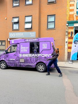 A purple van branded with 'Breakpoint' logos is parked on a city street while a person stands next to it, with a brick building behind them.