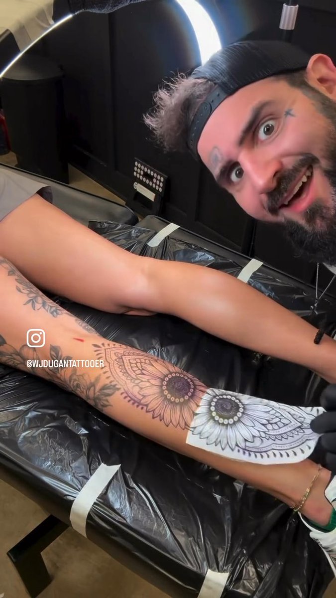 A tattoo artist wearing a black cap with a surprised expression in the background, while a detailed mandala tattoo is being applied to a client's leg in the foreground, tagged '@WJDUGANTATTOOER'.