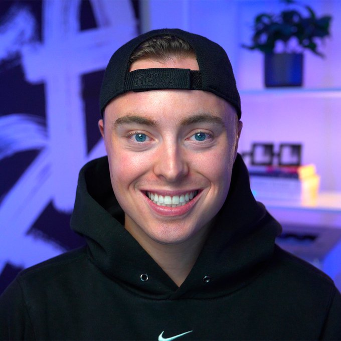 A close-up portrait of a smiling young man wearing a backwards black Toronto Blue Jays cap and a black Nike hoodie.