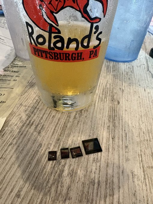 A nearly empty pint glass of beer branded 'Roland's Pittsburgh, PA' sits on a wooden table next to a receipt, with four SD memory cards arranged in a row.