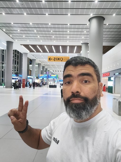 The image features a man with a beard wearing a white t-shirt who has made peace signs while standing in an airport terminal. The caption of the photo reads, "I'm in Istanbul, Turkey, waiting for my flight to Madrid; I’ll be in Madrid tomorrow, and there are only a few days left until the Solana Summit Berlin by @SuperteamDE." This post seems to be related to travel or an event happening in Madrid. The man's pose with peace signs suggests that he is excited about his upcoming trip or perhaps attending an important event like the Solana Summit Berlin.